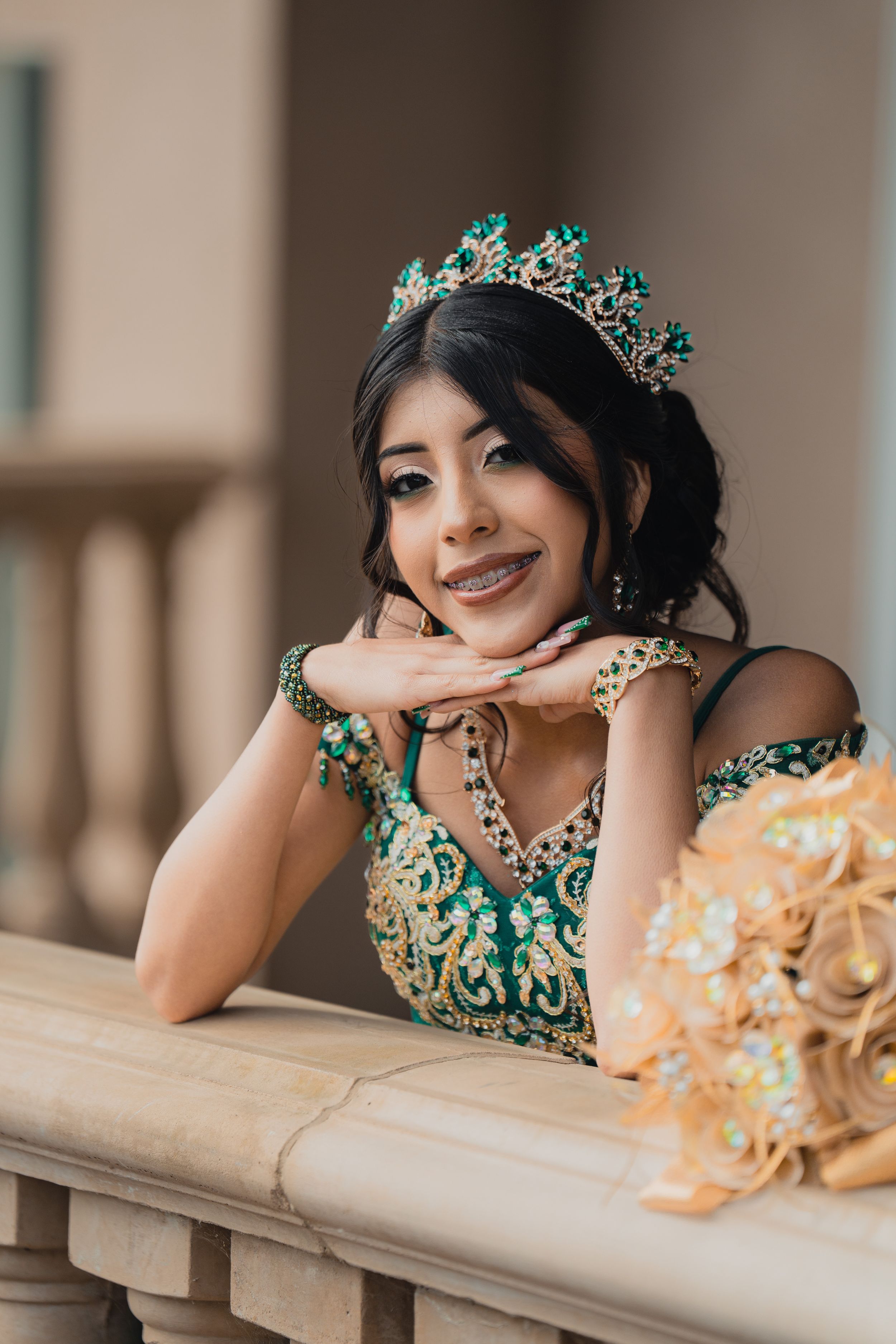 A person in an elegant emerald green gown and tiara poses gracefully along a beige stone balcony in a series of portraits.