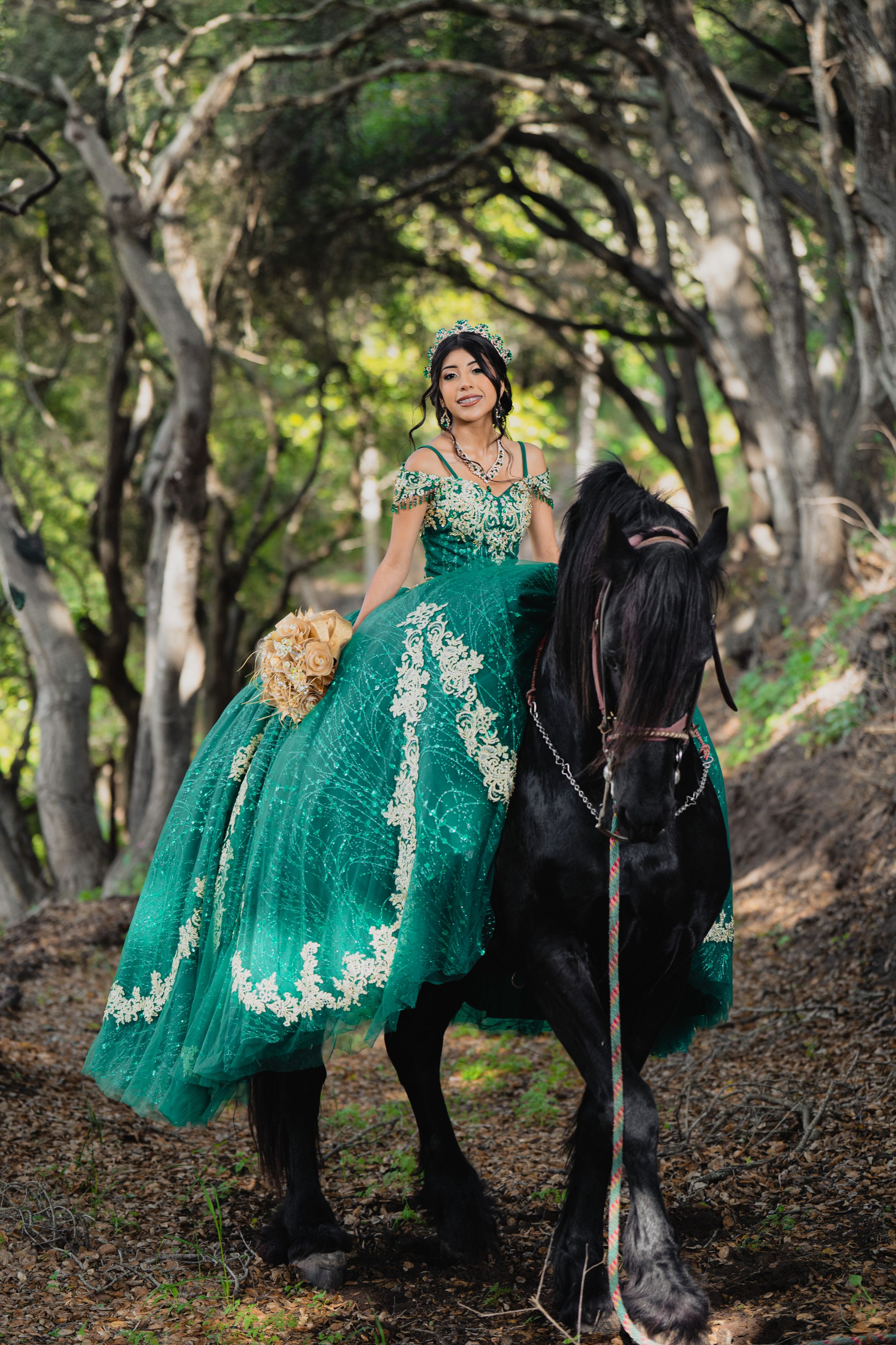 A figure in an emerald green and lace quinceañera dress poses elegantly atop a black horse in a tree-lined path.