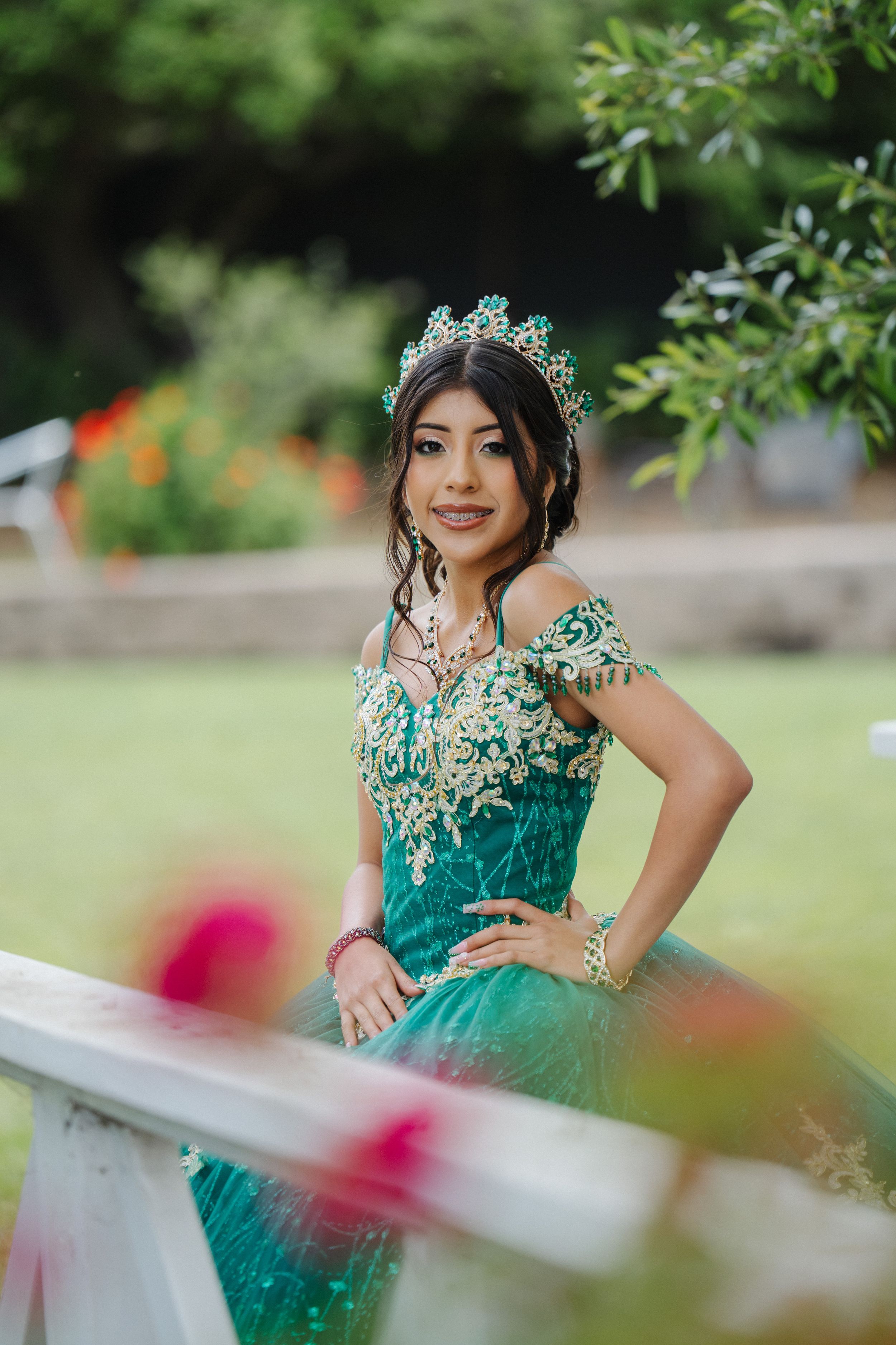 A person in an elegant teal gown with lace details and a crown poses in a garden setting with blurred flowers.