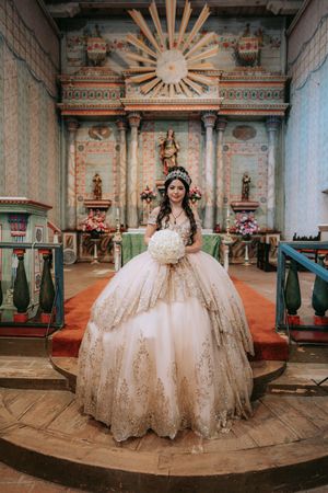 Ornate white and gold quinceañera ball gown photographed in a historic church with decorative sunburst architecture.
