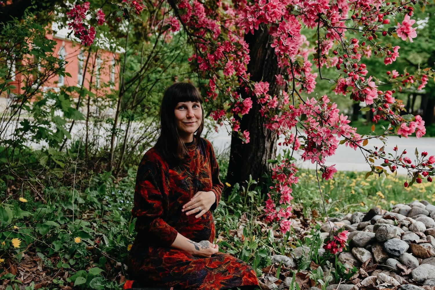 A woman in a red dress sits beneath blooming pink flowers in a tranquil garden setting.