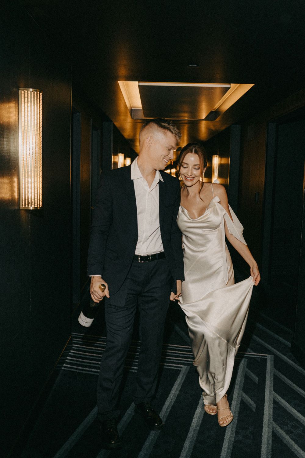 A couple in formal attire walk together intimately down a dimly lit hotel hallway with elegant wall sconces.