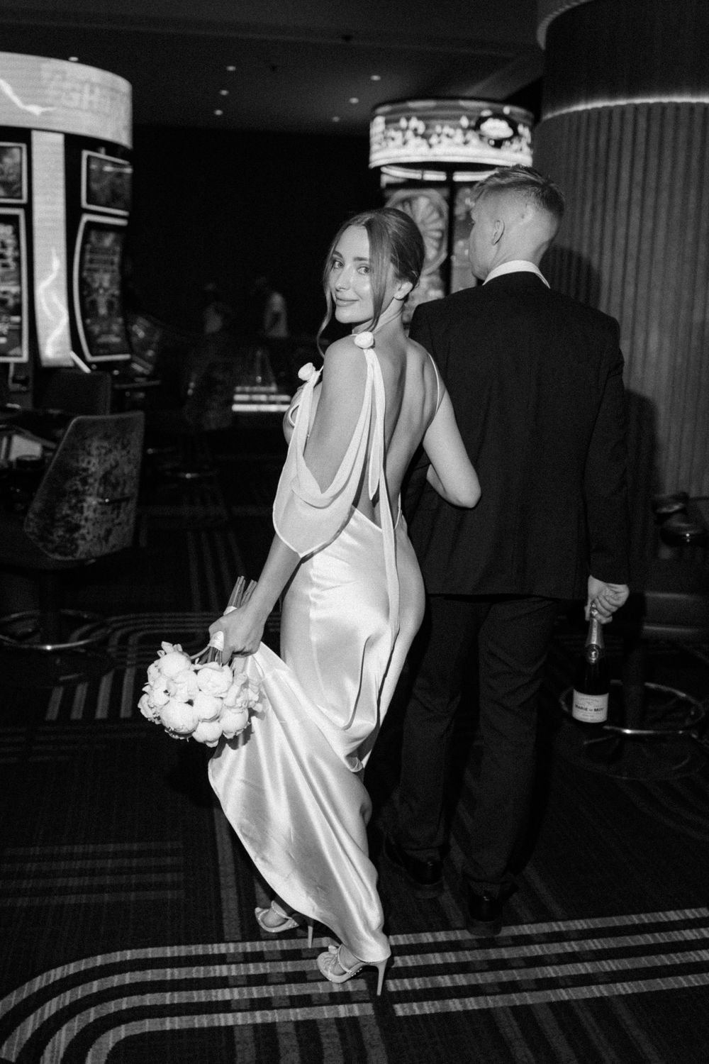 Black and white wedding photo showing bride in sleek backless satin gown holding bouquet at evening reception.