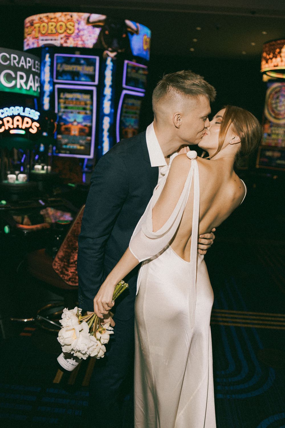 A bride and groom share a romantic kiss in a casino with slot machines and neon lights in the background.