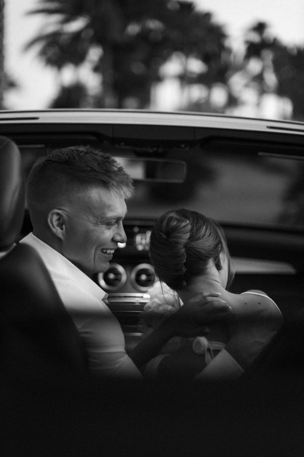 Black and white candid photo showing two people in formal attire sitting in a car together, shot through the window.
