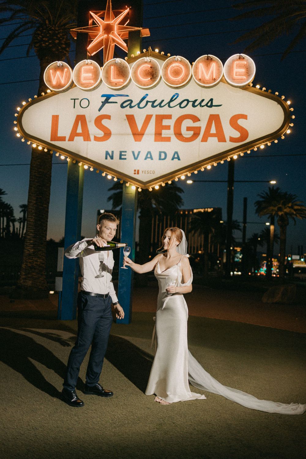 A newlywed couple poses playfully in front of the iconic Welcome to Las Vegas sign at night.