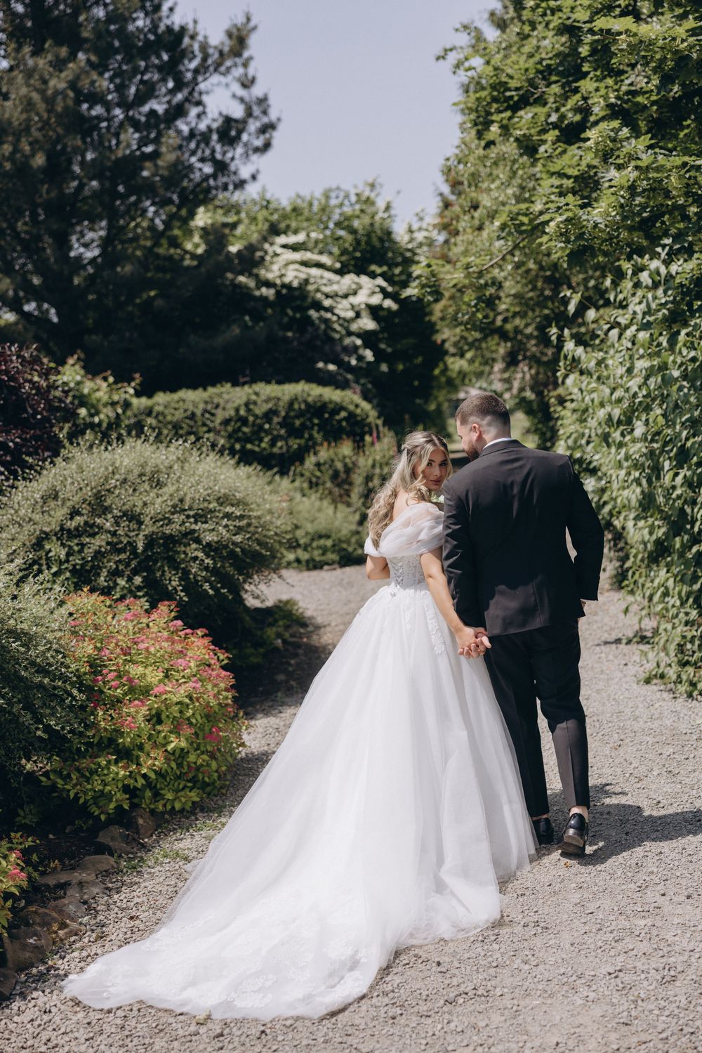 A wedding couple walks along a gravel path surrounded by lush greenery and blooming flowers in an outdoor garden setting.