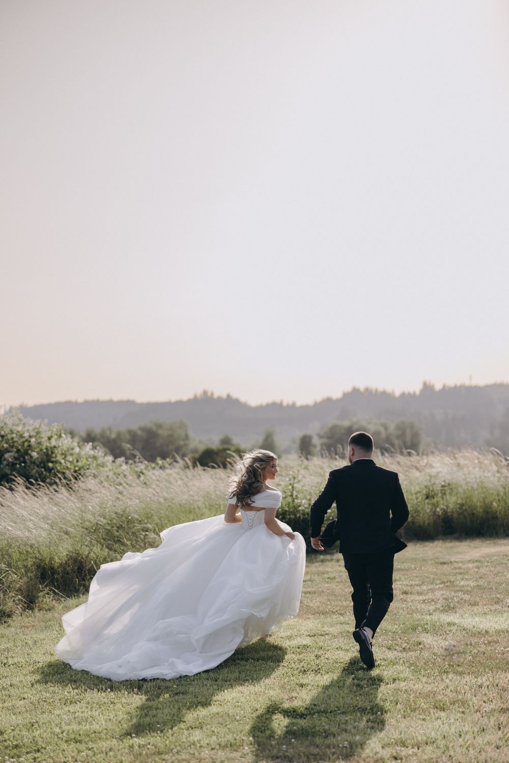 A couple running through a meadow of tall grass with the bride's white dress flowing in the wind against a hazy landscape.