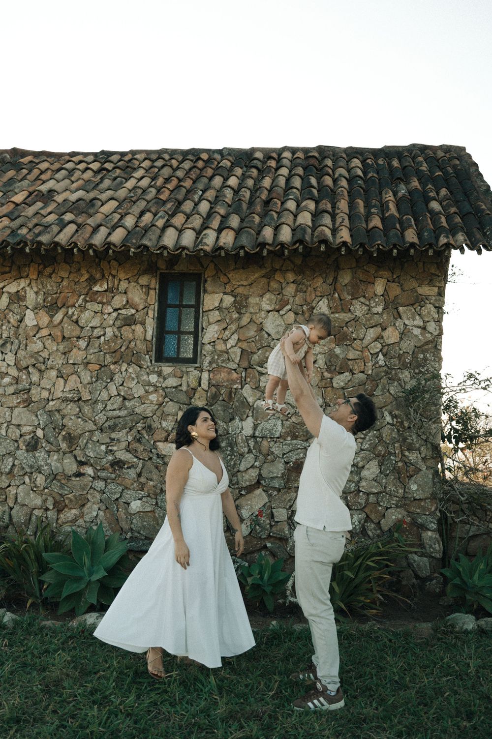 A couple in white outfits playing with their baby against a rustic stone house backdrop.