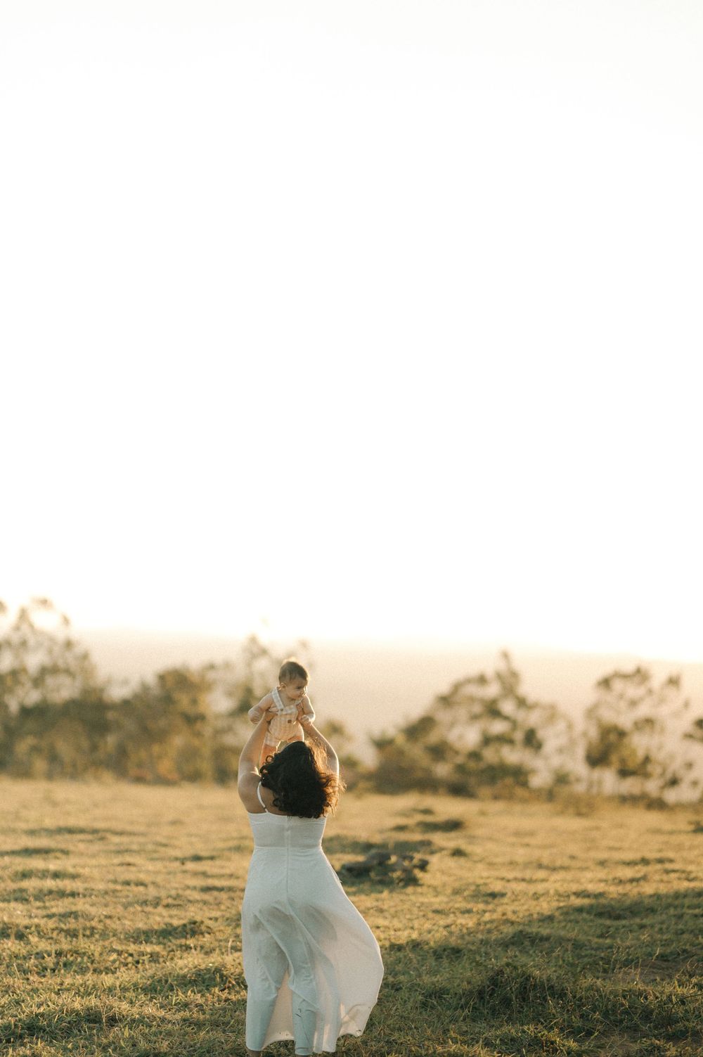 A person in a flowing white dress stands in a sunlit meadow at golden hour with trees silhouetted in the background.