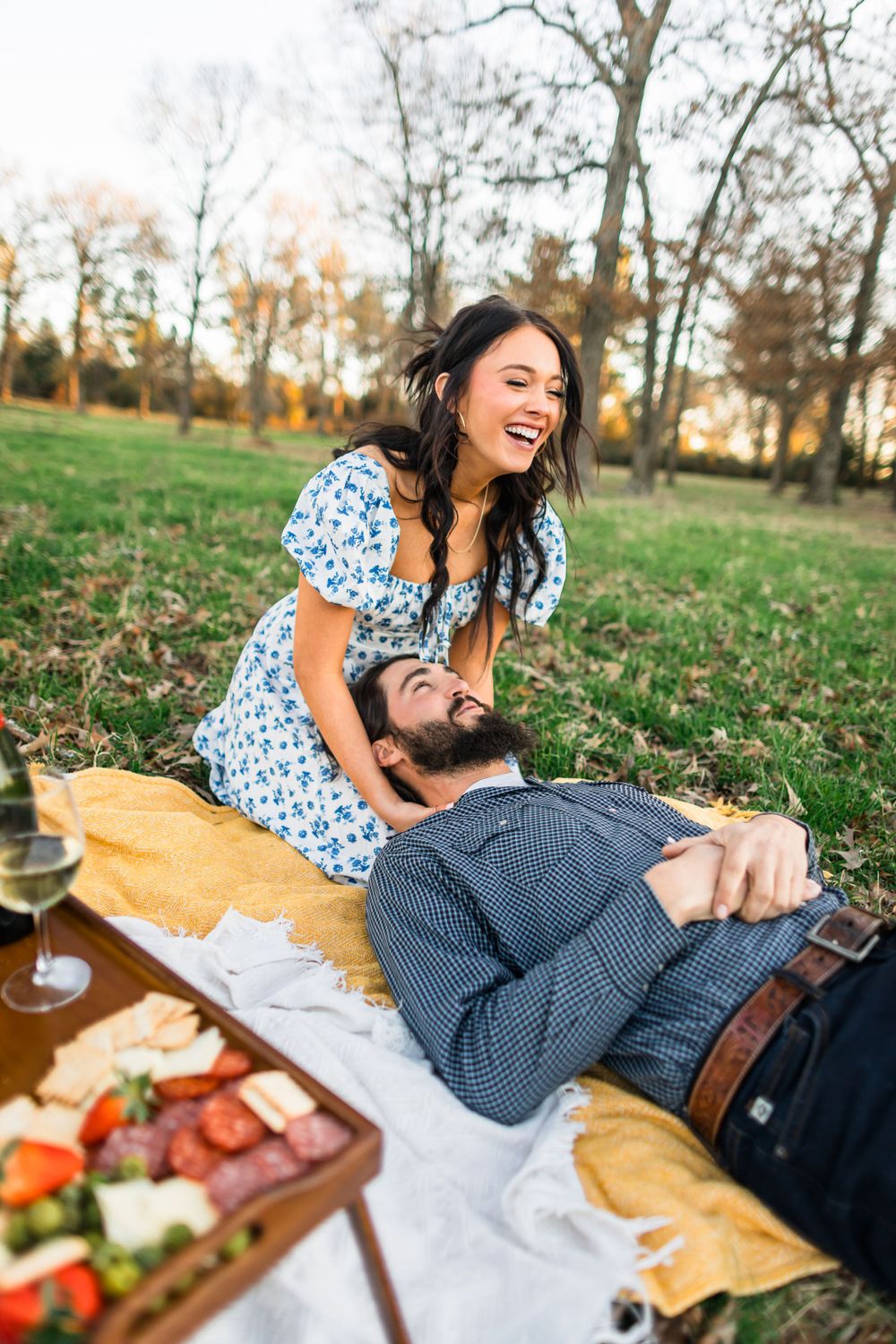 A playful outdoor picnic scene with a couple laughing on a yellow blanket with food and drinks.