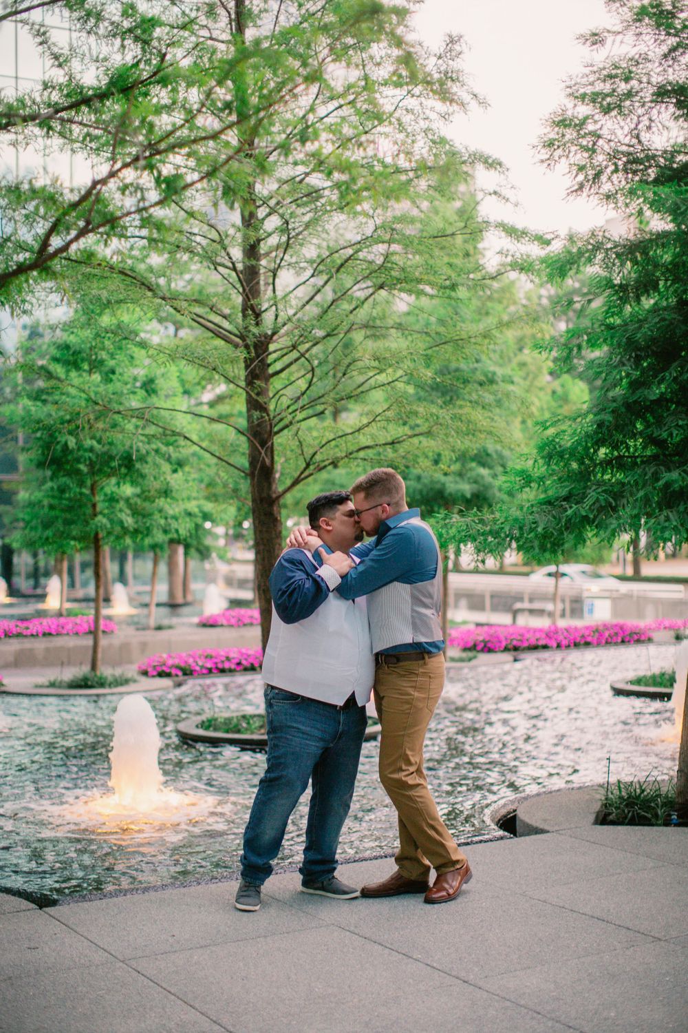 Tender embrace near illuminated fountain with pink flowers and trees in background.