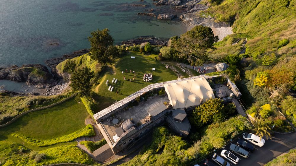 Aerial view of a coastal property with lush green grounds, stone building, and rocky shoreline in soft golden light.