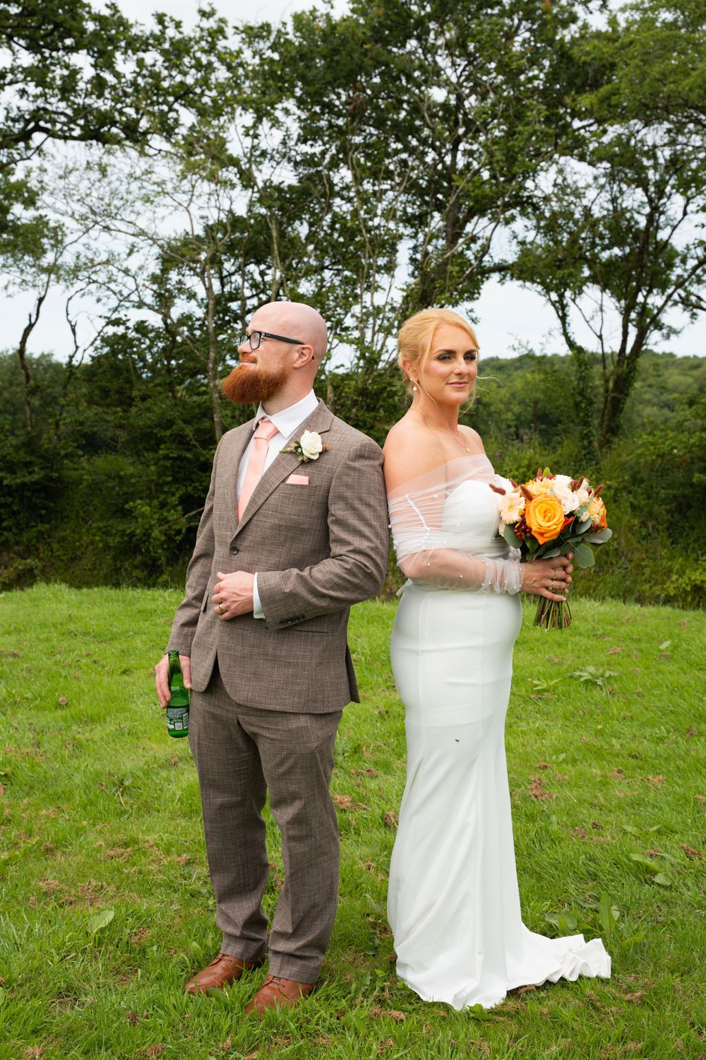 Bride in white gown holding orange bouquet stands back-to-back with groom in brown tweed suit on green lawn.