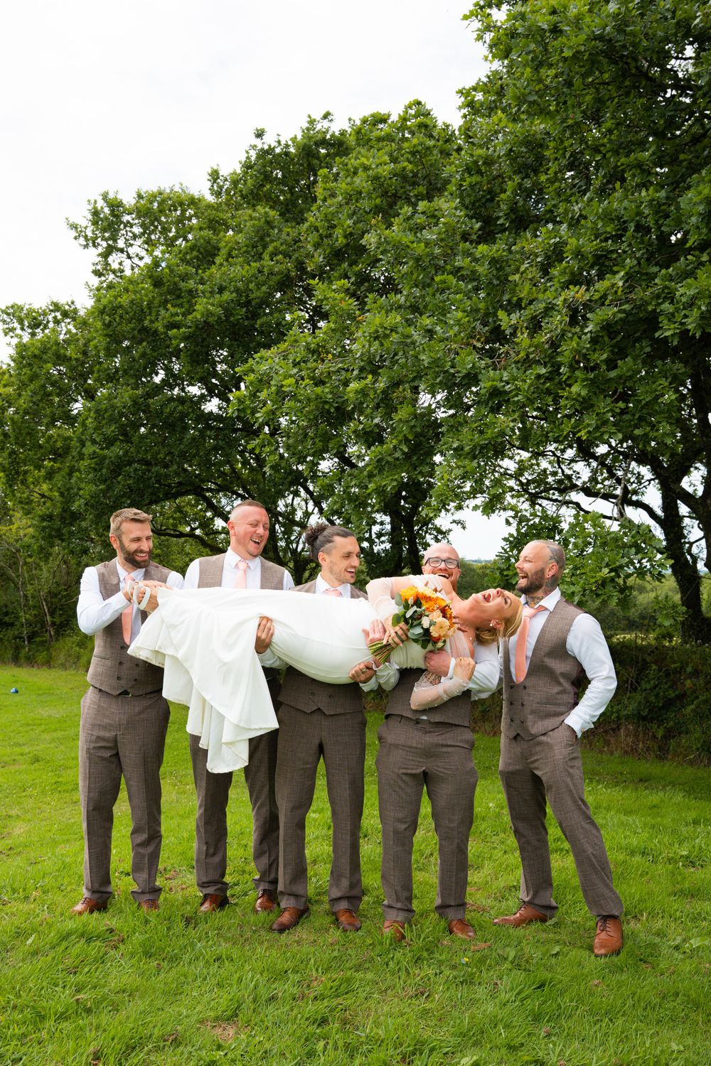 Groomsmen in brown suits carry a laughing bride in a white dress outdoors on green grass with trees behind.