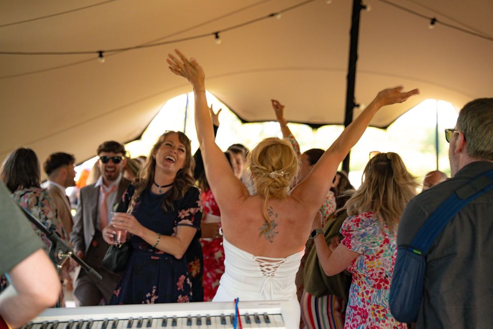 Bride with tattoos raises arms joyfully on wedding dance floor under a tent with guests celebrating around her.