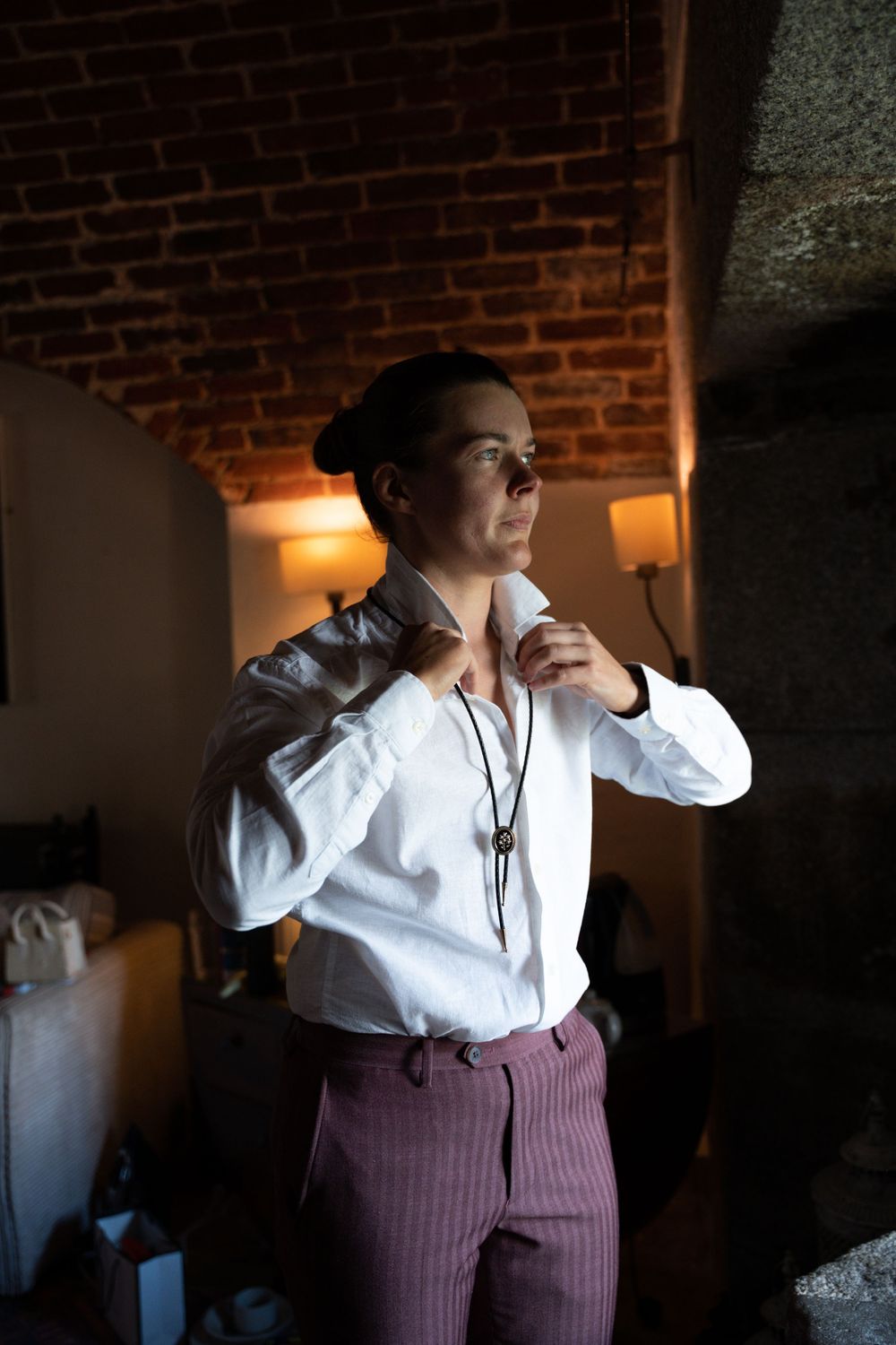Person in white shirt and striped pants adjusting collar in dimly lit room with brick ceiling.