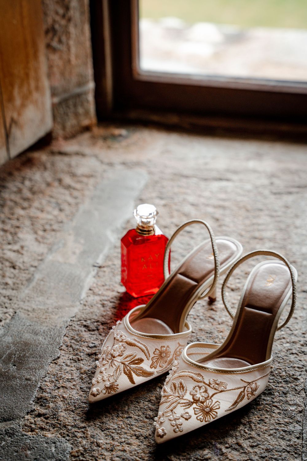 Elegant white embroidered bridal heels beside a red perfume bottle on stone floor near a window.