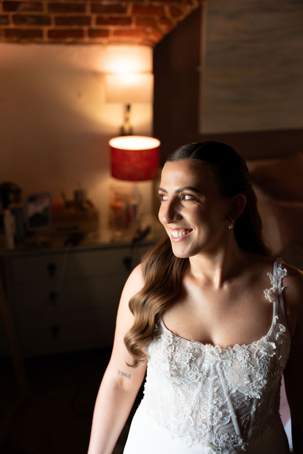 Smiling bride in lace wedding dress standing in warmly lit room with brick wall and glowing lamp.