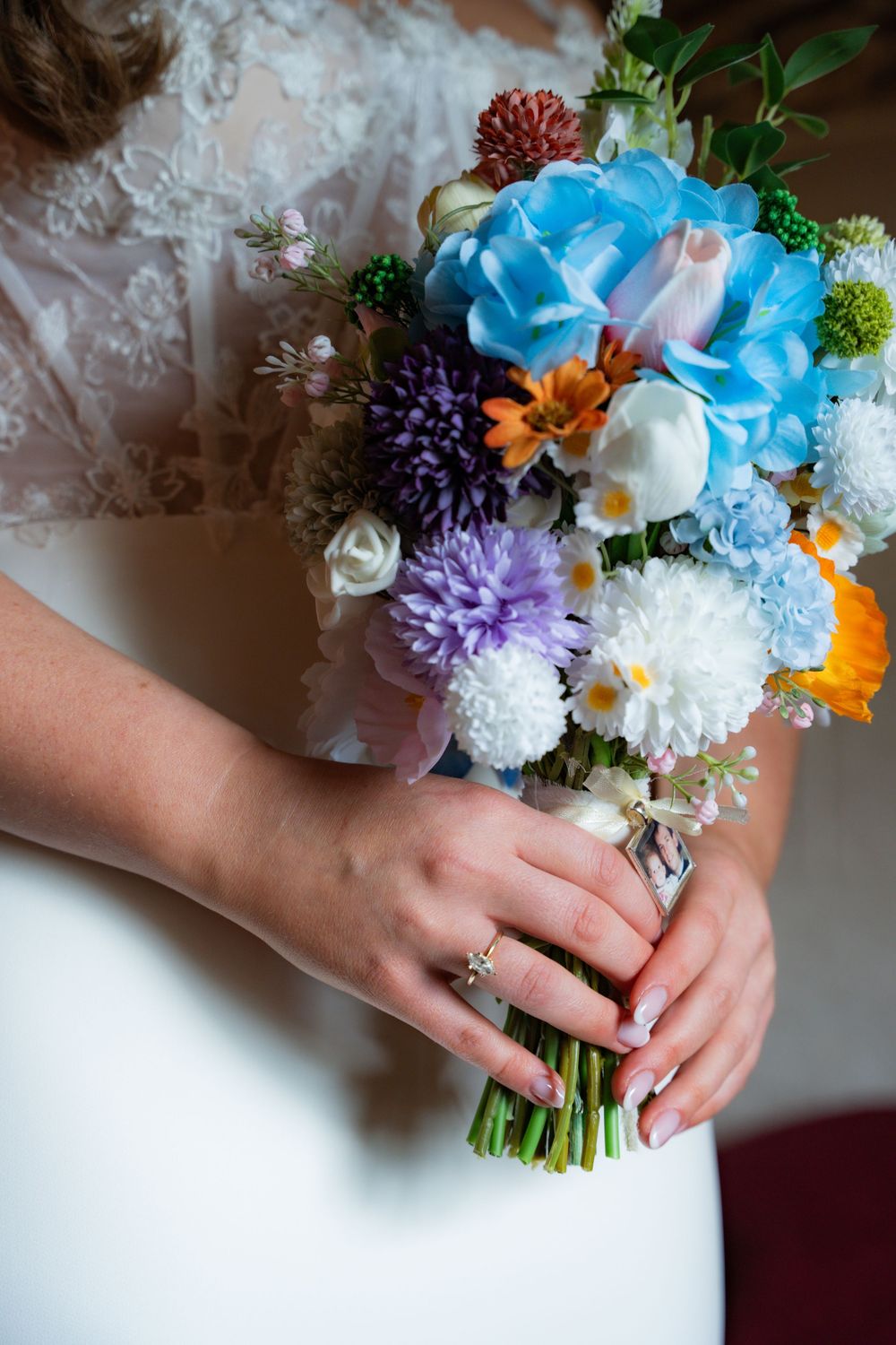 Bride holding a colorful wedding bouquet with blue, purple, white, and orange flowers, wearing a diamond ring on her finger.
