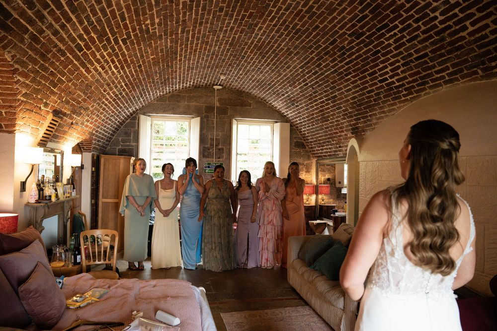 Bride in white gown faces bridesmaids in colorful dresses inside a rustic brick vaulted ceiling venue.
