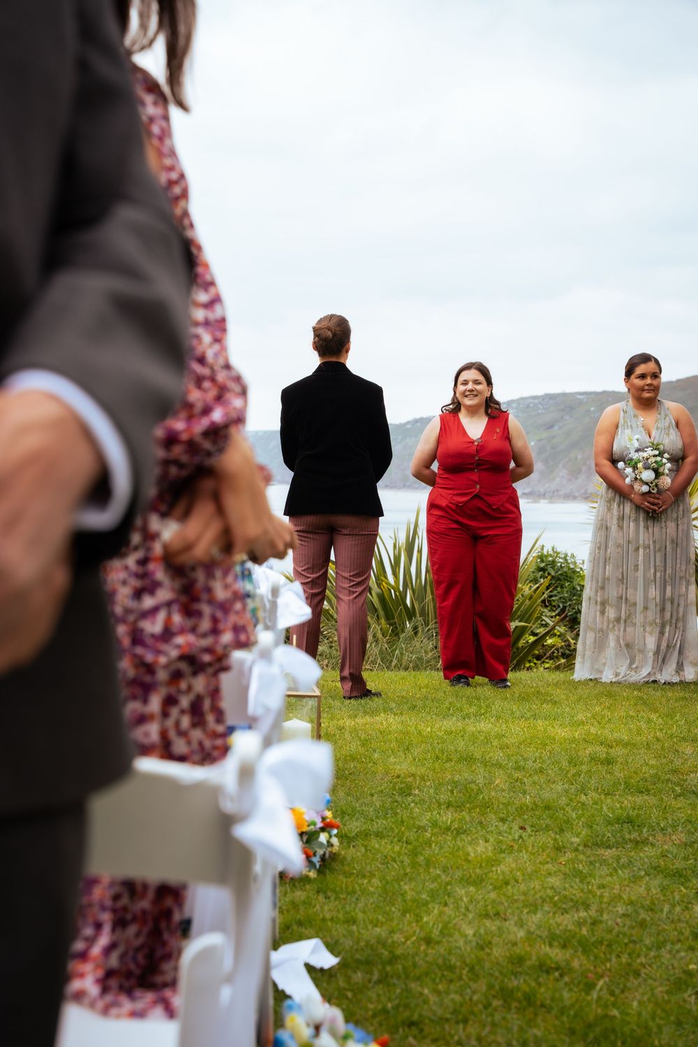 Outdoor wedding ceremony on green lawn with bridal party in red and gray dresses, white chair aisle, coastal cliffs backdrop.