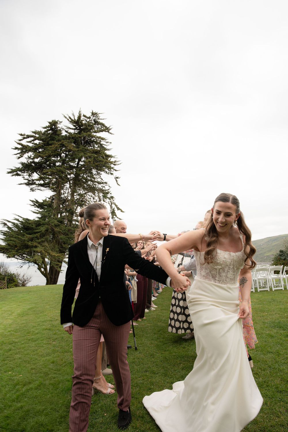 Two brides walk hand-in-hand down the aisle at an outdoor wedding ceremony on a green lawn.