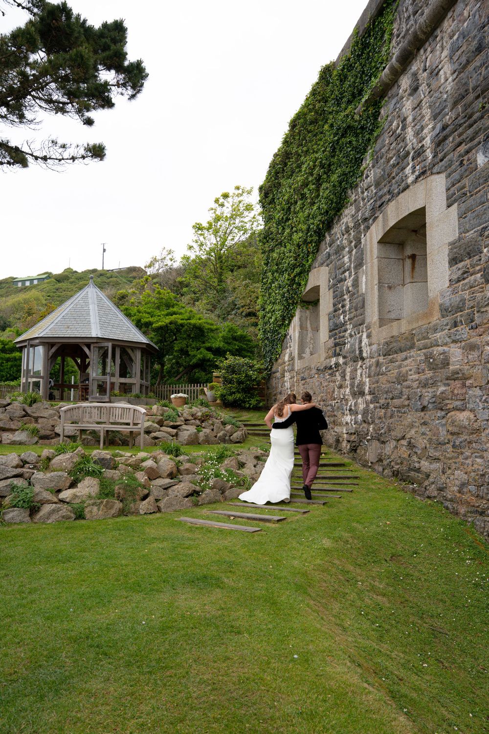 Bride and groom walking hand in hand along stone castle wall with ivy, garden gazebo visible in background.