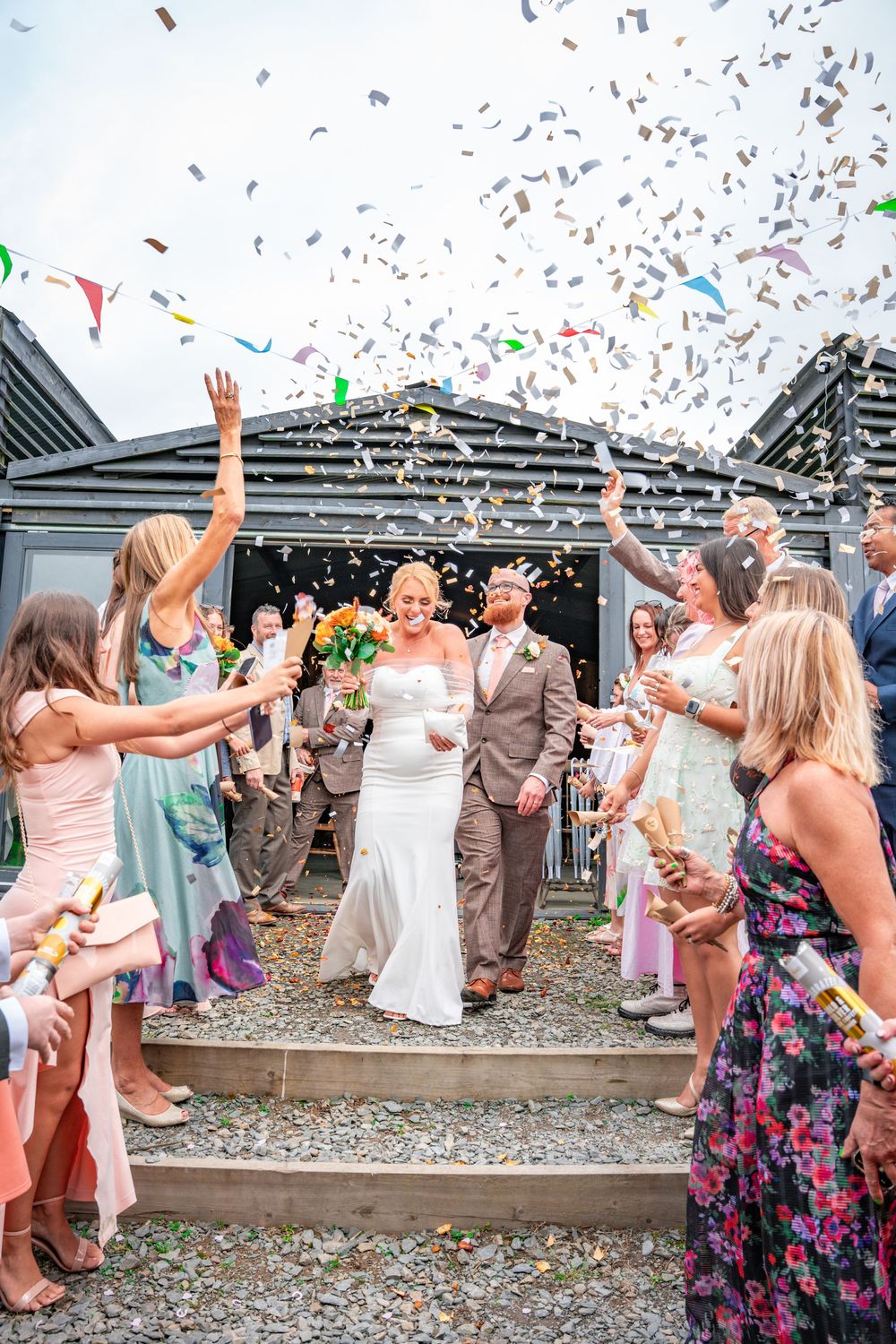 Bride and groom exit wedding venue through confetti shower, guests celebrating outdoors on sunny day.