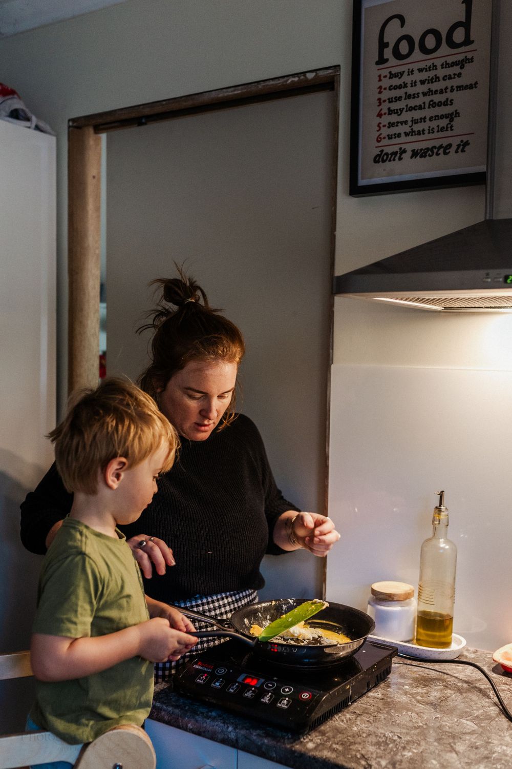 A mother stands in her kitchen cooking eggs, with a toddler standing on a step stool working alongside her.