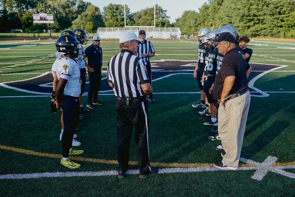 A referee in black and white stripes signals from the football sideline during a game.