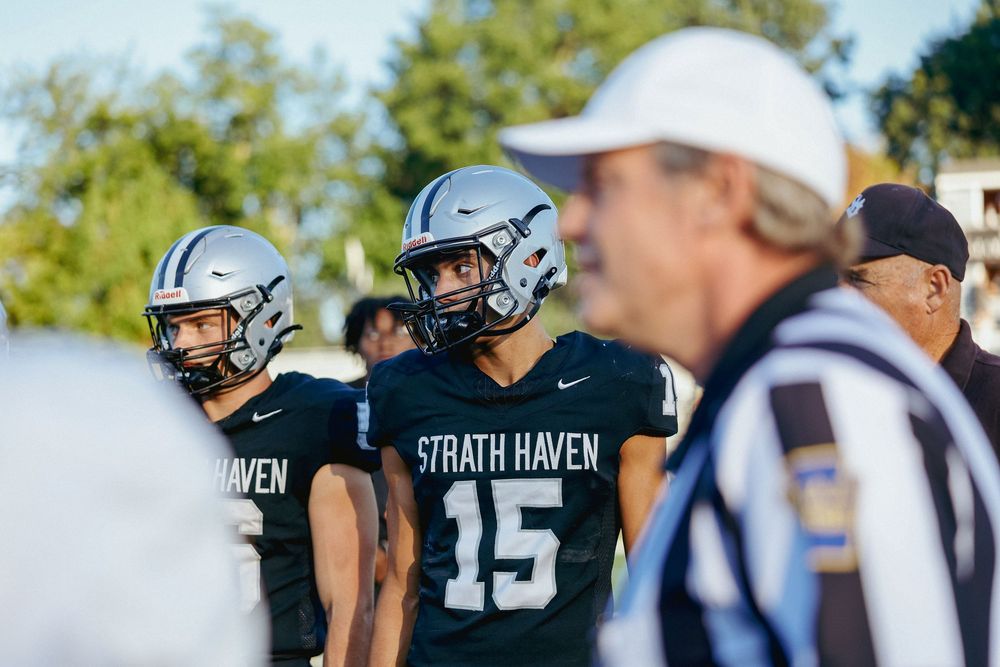 High school football players huddle on the sideline during a game wearing black and silver uniforms with coaches nearby.