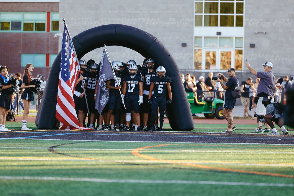 American football players line up in matching uniforms during a night game under stadium lights and inflatable arch.