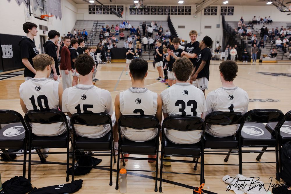 Basketball players and coaches sit on sideline benches during a game in a high school gymnasium.