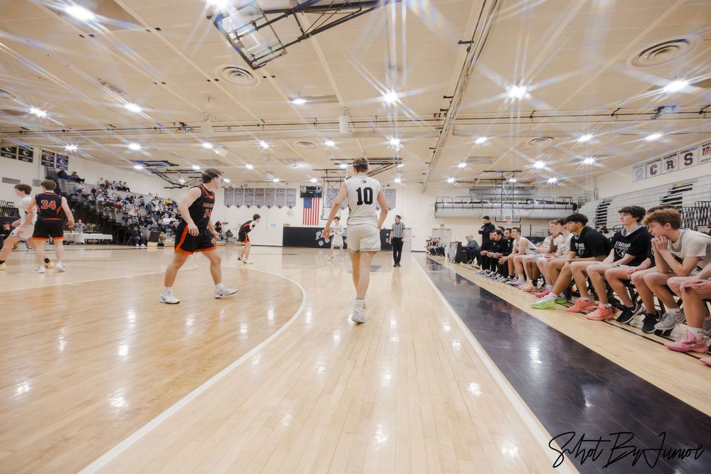 Basketball players and spectators gather in a bright indoor gymnasium with polished wooden floors and fluorescent lighting.
