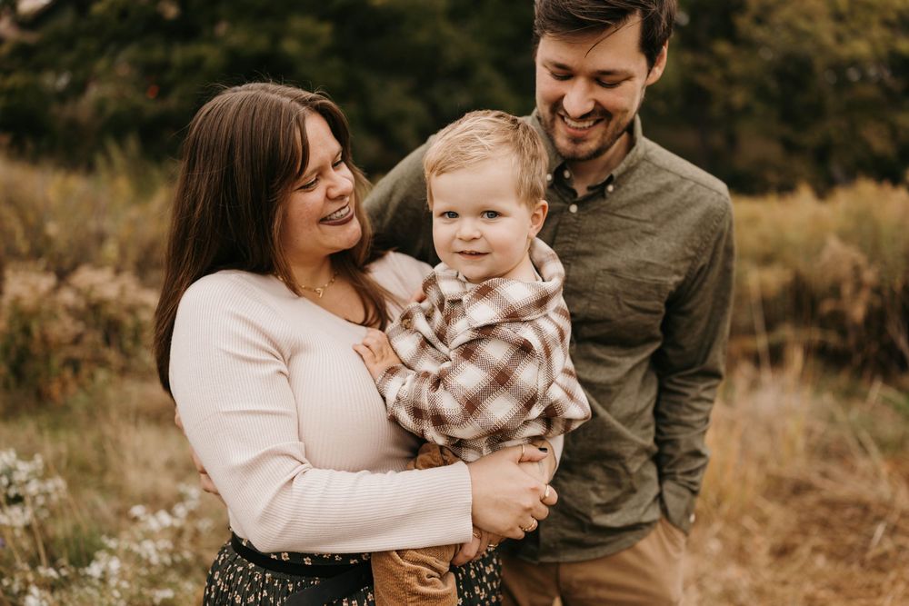 Cheerful family of three with toddler boy poses in autumn wildflower field.