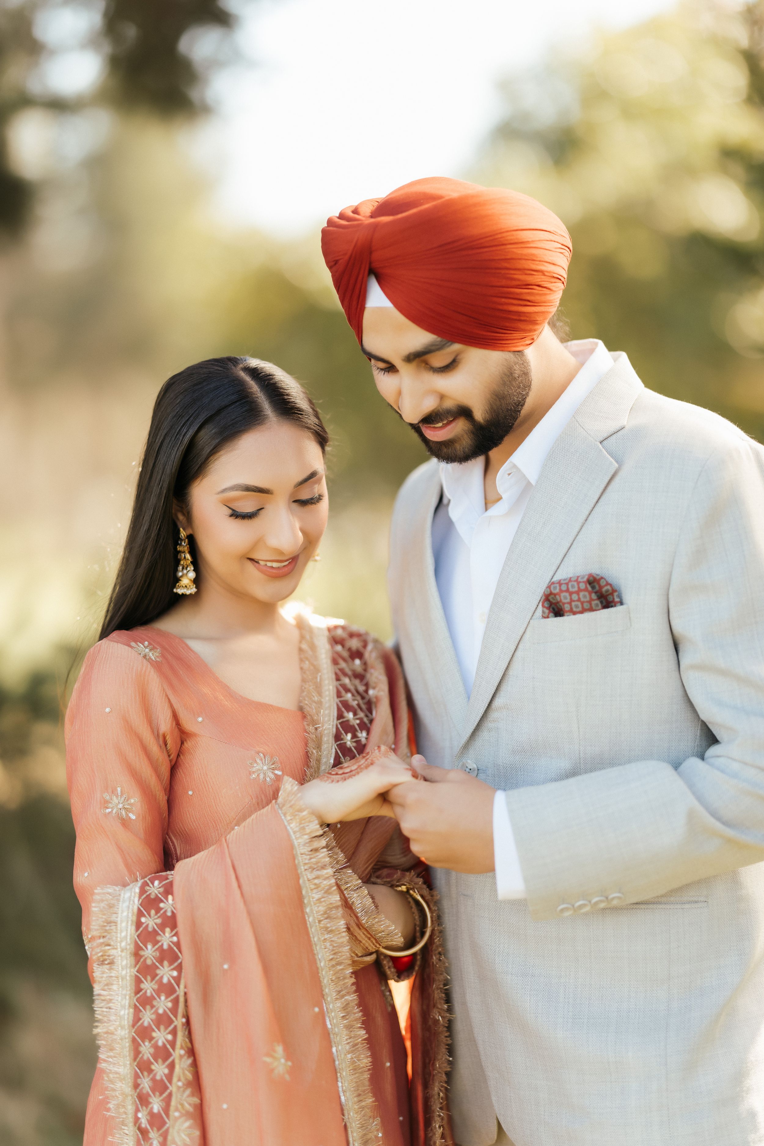 A couple in traditional Indian attire share an intimate moment outdoors, with the woman in peach and man in light gray suit.