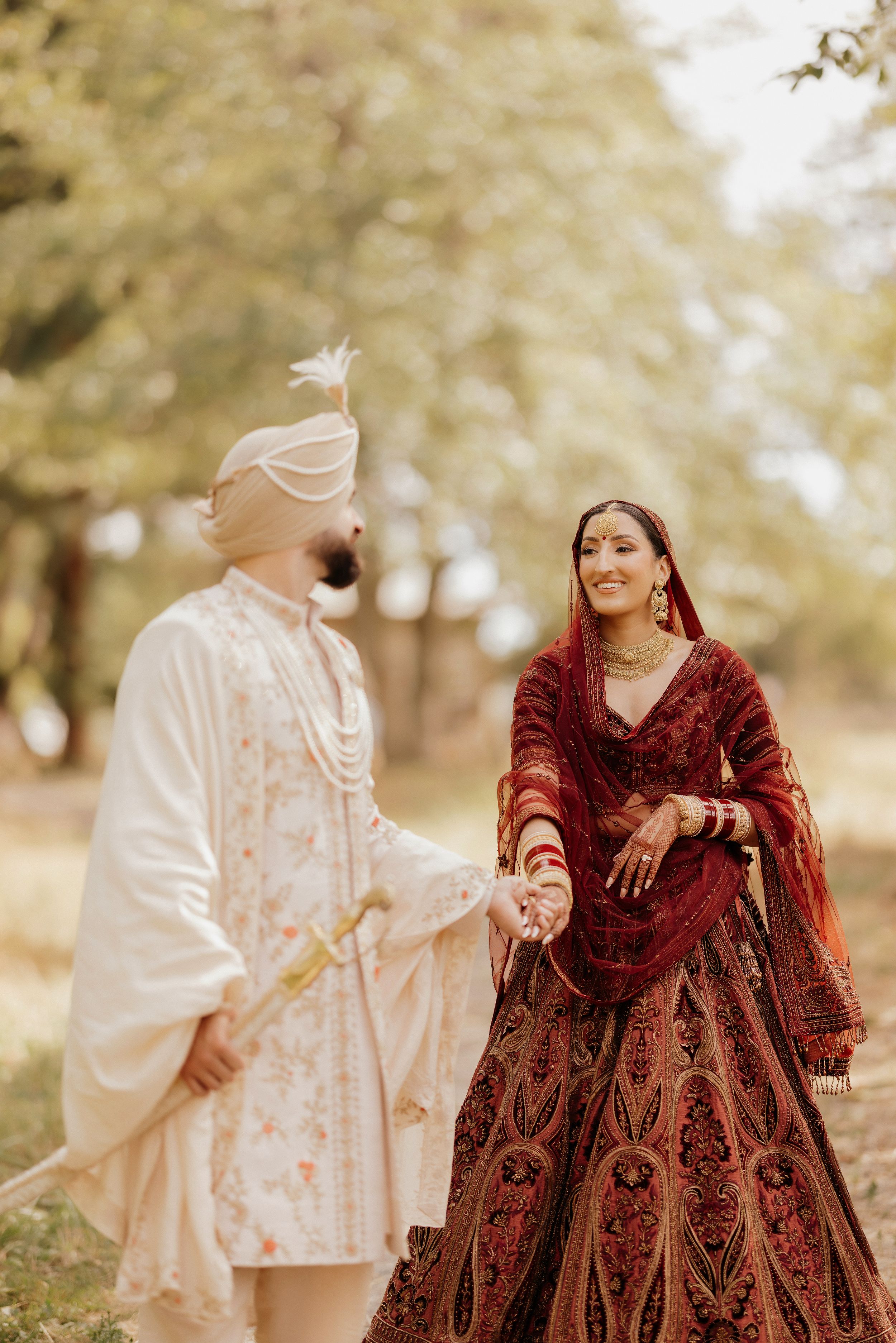 A couple in traditional Indian wedding attire walks through a dreamy outdoor setting in maroon and cream colors.