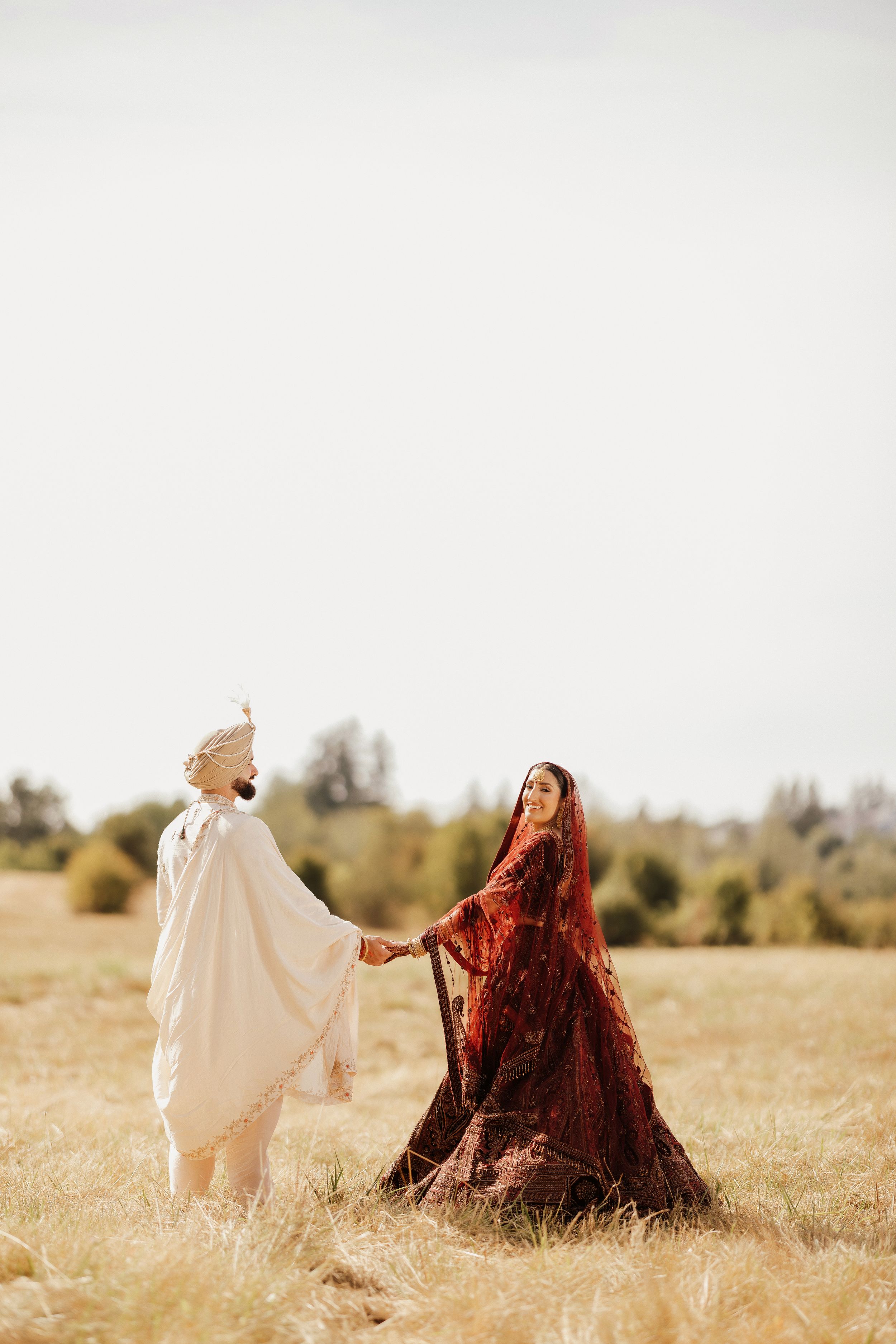 A couple in traditional attire sits in a golden field during sunset, creating a romantic and ethereal atmosphere.