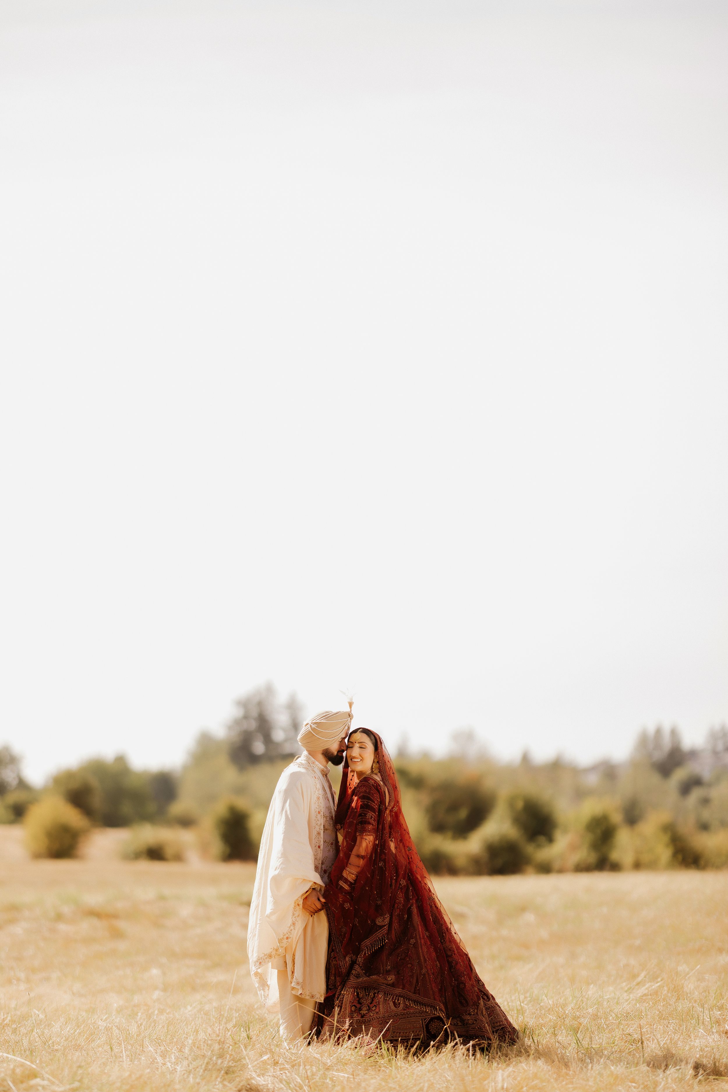 A couple in traditional South Asian wedding attire embraces in a golden field during sunset.