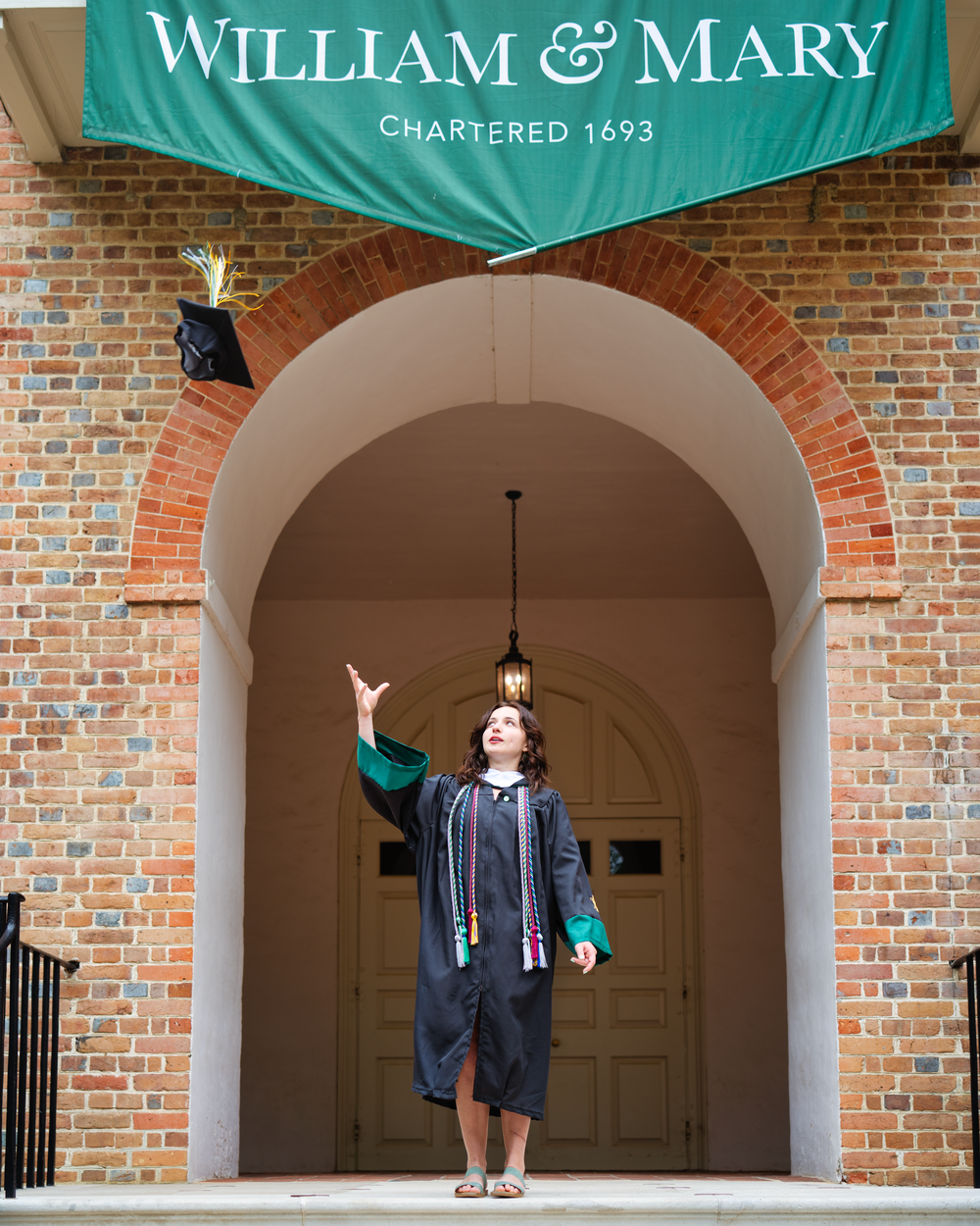 Graduate posing with celebration cap toss under William & Mary archway entrance with teal banner.