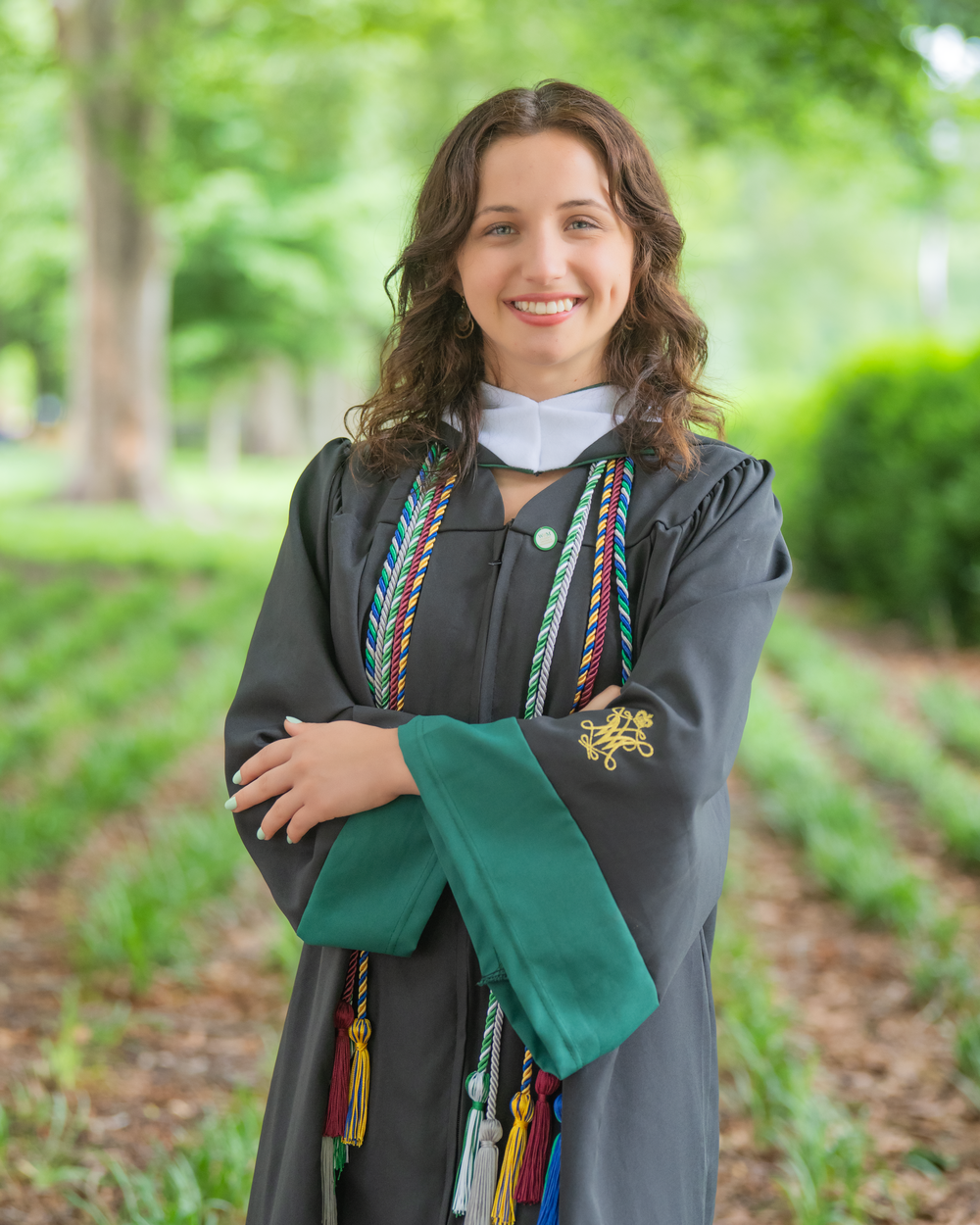 Graduate in commencement regalia with decorative cords and tassels poses outdoors on a spring day.