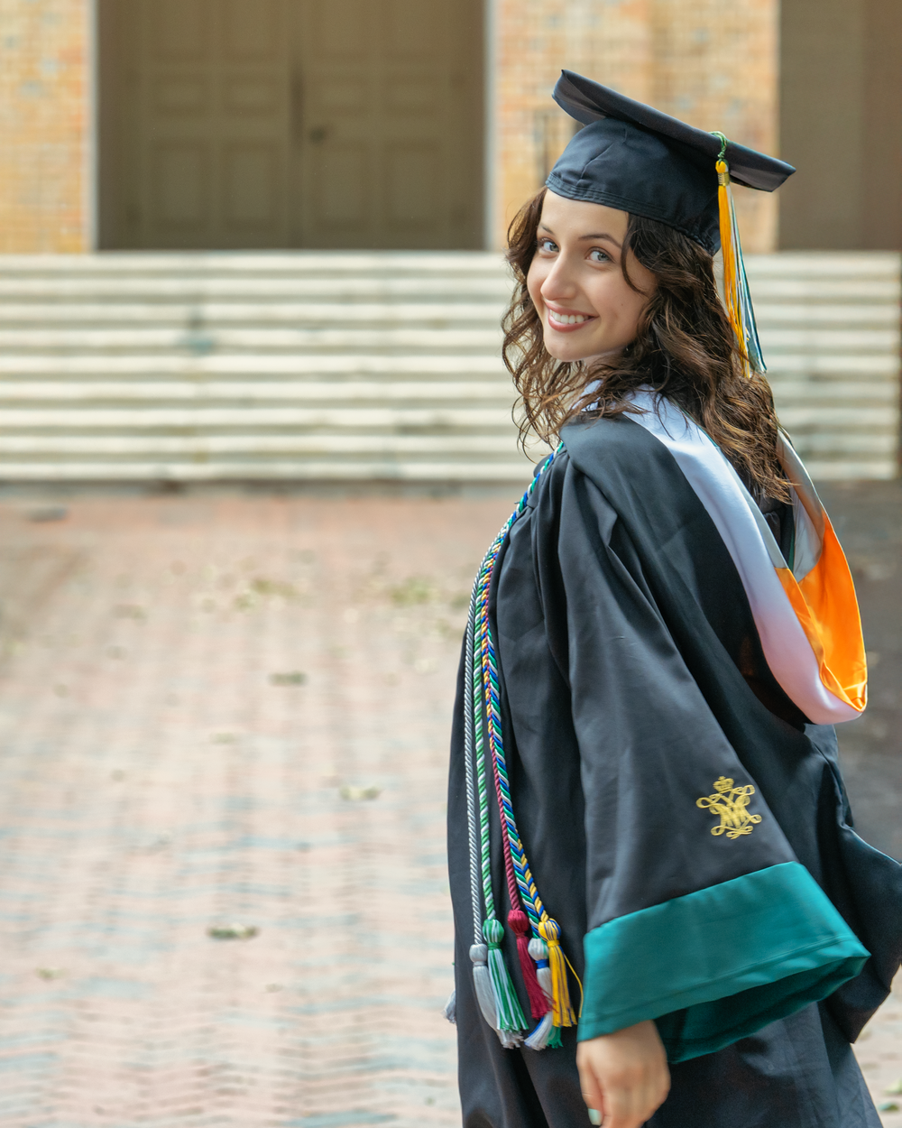 Graduate in cap and gown with graduation cords smiling while standing on brick walkway outside academic building.