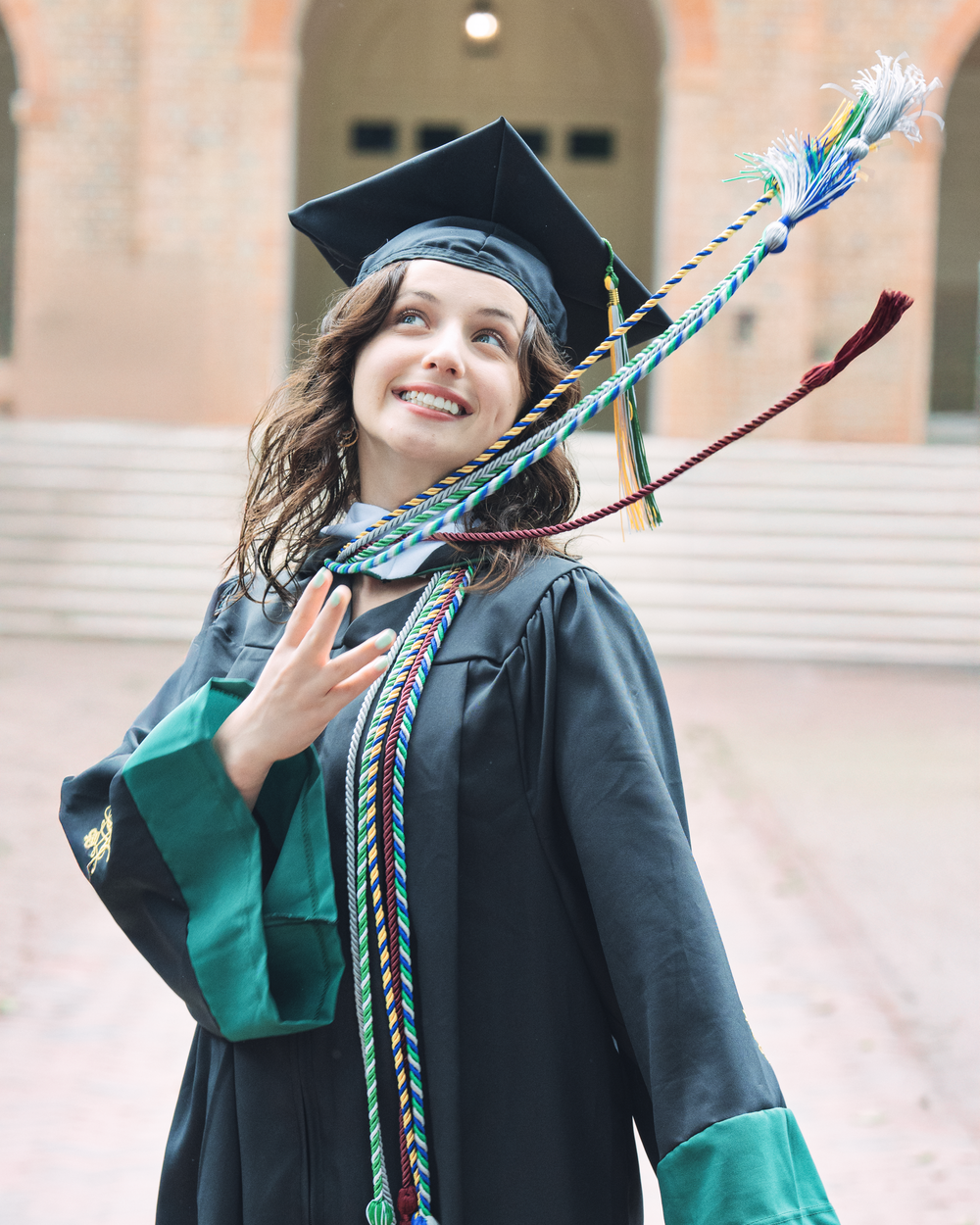Graduate in cap and gown holding decorative graduation cords poses joyfully on campus steps outside.