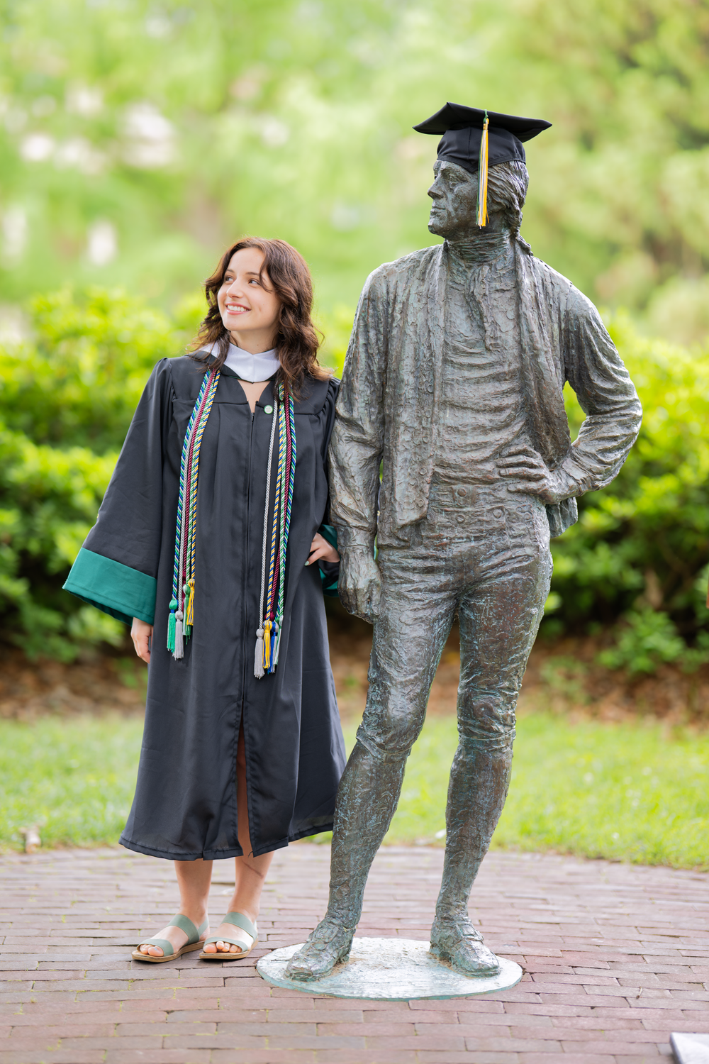 Graduate in academic regalia poses with a life-sized metallic statue wearing a graduation cap on a brick path.