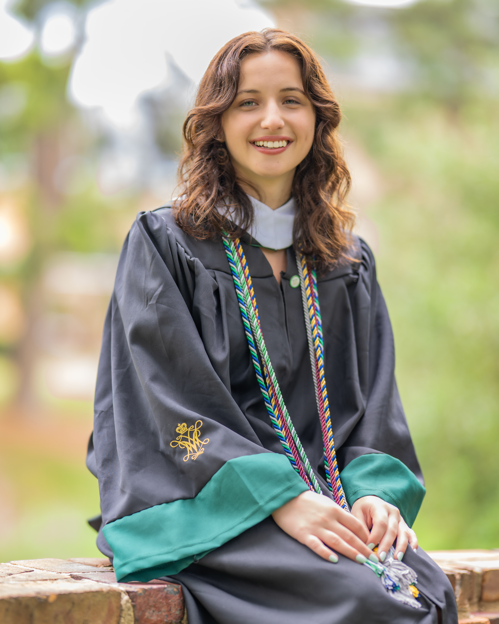 Graduate wearing a black academic gown with green accents and colorful cords poses outdoors for a graduation photo.