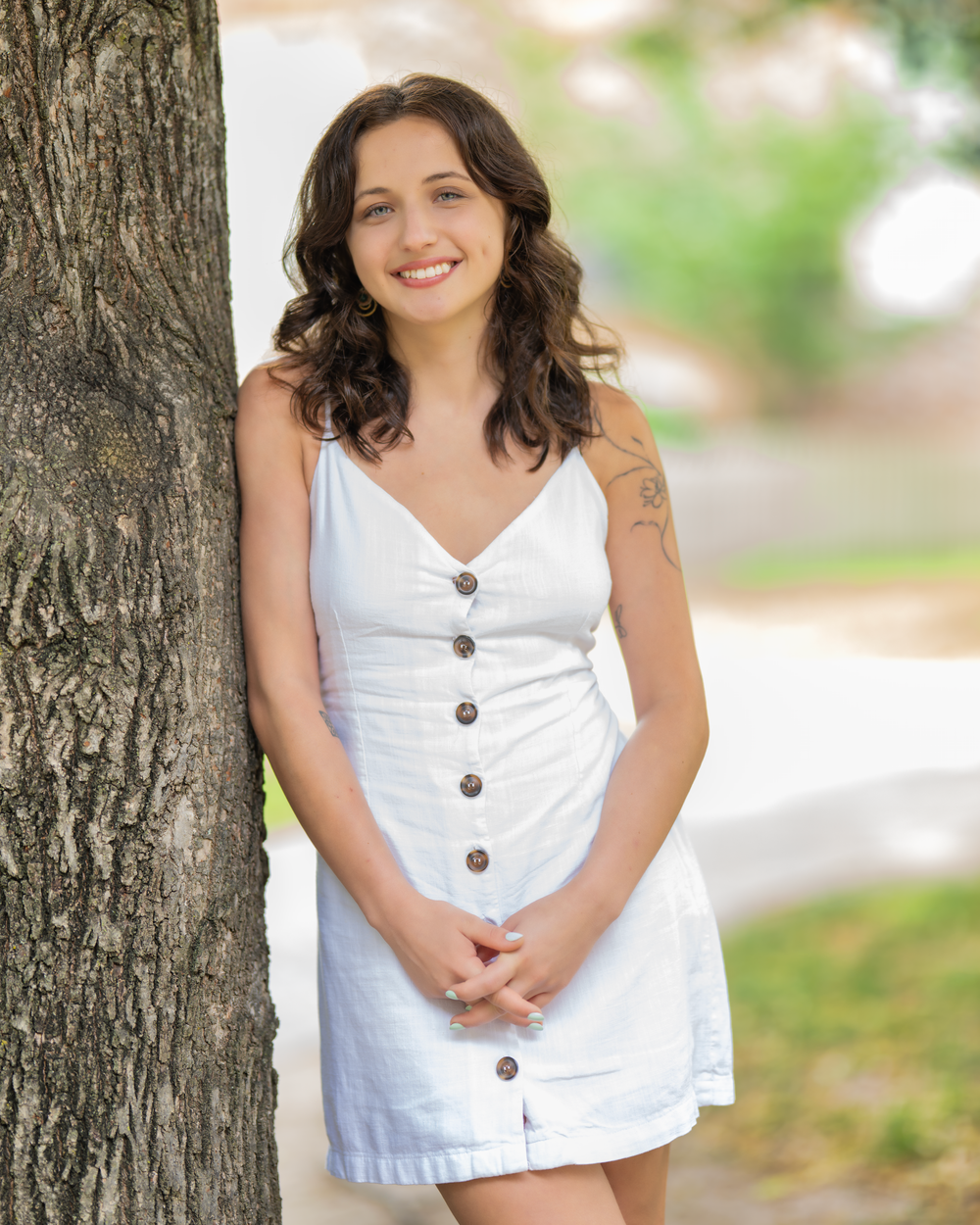 Woman in white button-up sundress poses against tree trunk on sunny day.