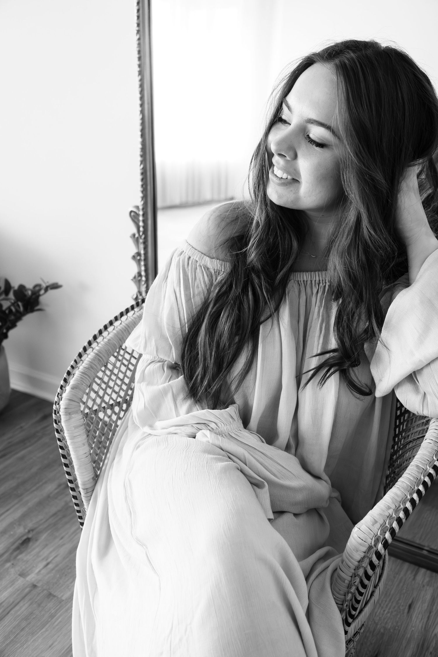 Black and white timeless portrait of someone relaxing in a wicker chair wearing a flowing white dress with long dark hair. Rochester NY