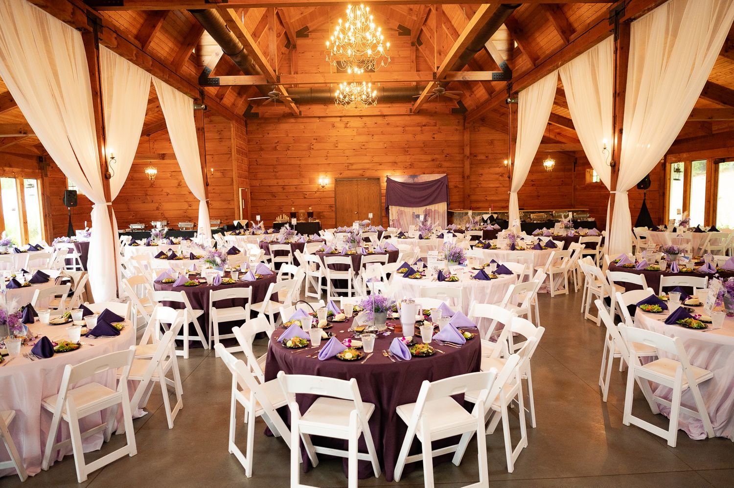 An elegant wedding reception venue with white chairs and purple decor in a rustic wooden barn.
