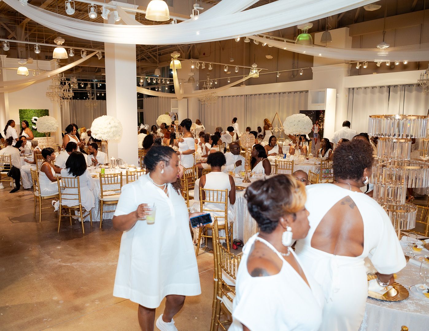 People in white attire mingle at tables in an elegant event space with industrial decor.
